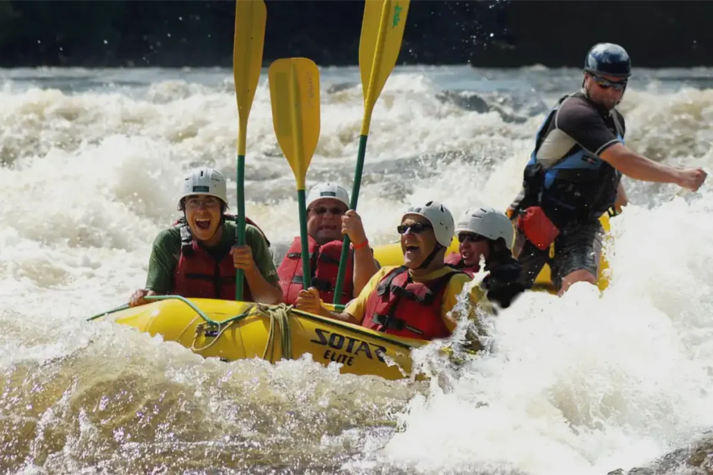 Five people in helmets and life jackets raft through whitewater in a bright yellow inflatable boat, guided by a standing instructor at the back.